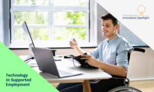 Person in a wheelchair that is smiling and looking at a computer while jotting down notes in a notebook. The overlay in the bottom left corner is a green triangle and the words "Technology in Supported Employment". In the top right corner, there is a grey overlay with the Innovation Spotlight logo over top.